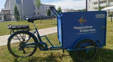 Picture of blue bike with carrying space in front, with the logo "Neighbourhood Concierge", in background neighbourhood residential area with big green space