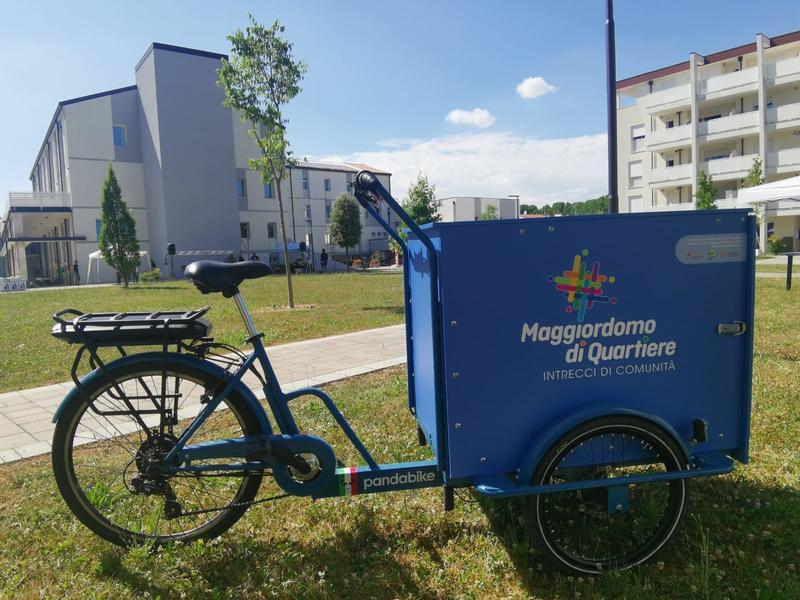 Picture of blue bike with carrying space in front, with the logo "Neighbourhood Concierge", in background neighbourhood residential area with big green space